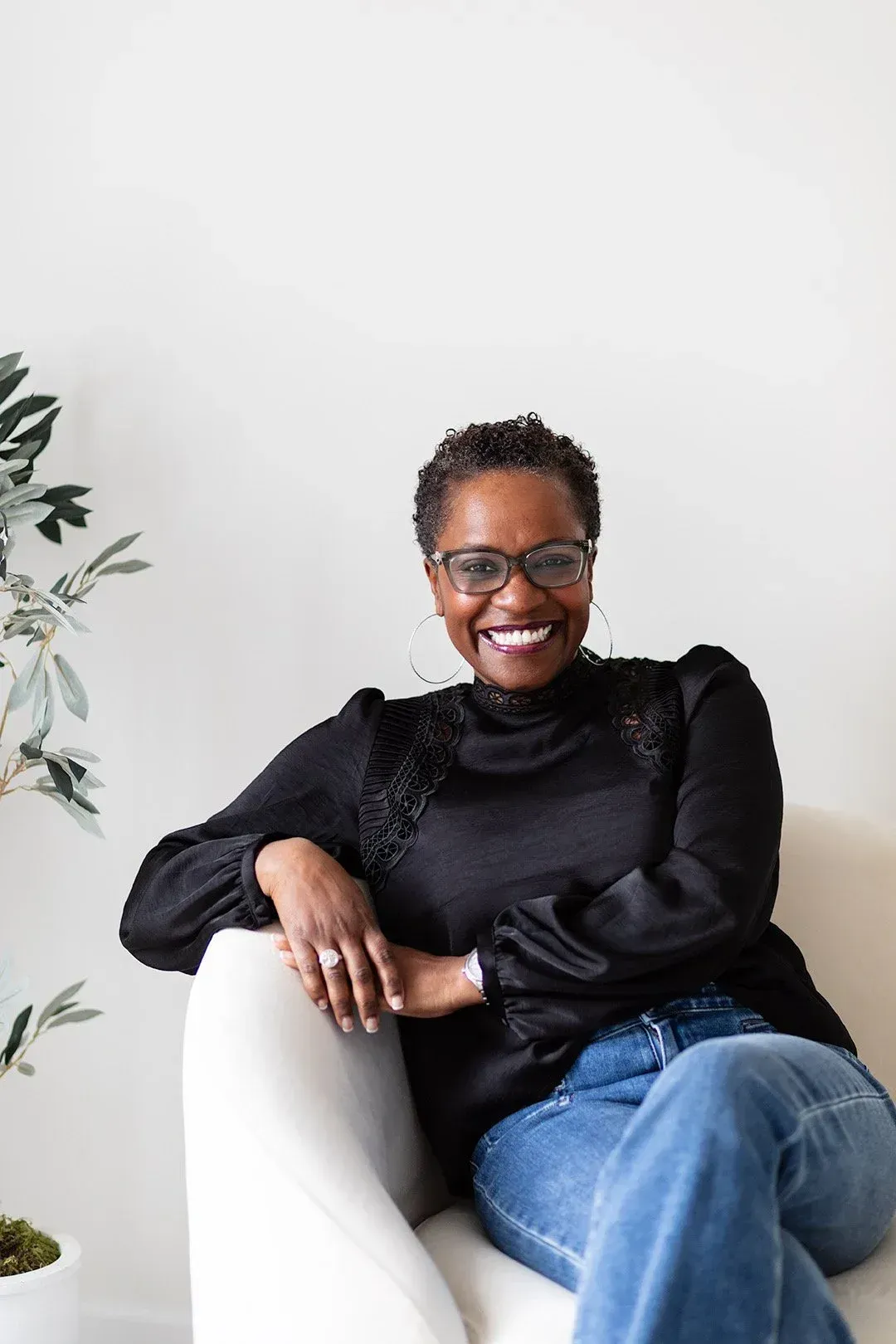Smiling person in black top and jeans seated on a white chair beside a potted plant