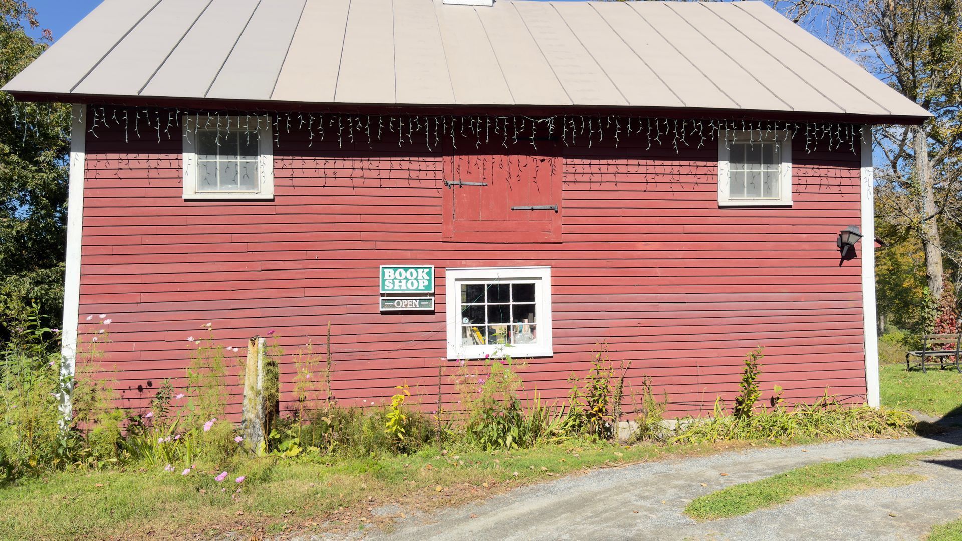 Red wooden barn with a metal roof and small windows beside a gravel path and greenery.
