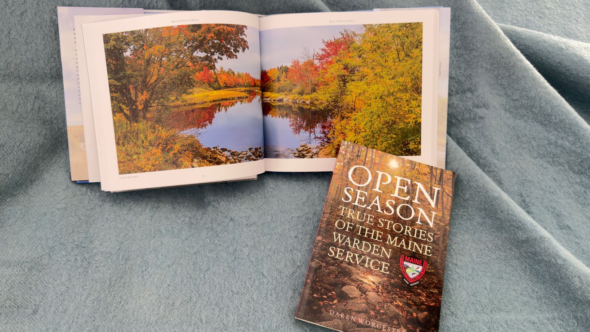 Open book on blue fabric beside a nature booklet with autumn trees and a stream. The book is called Open Season.