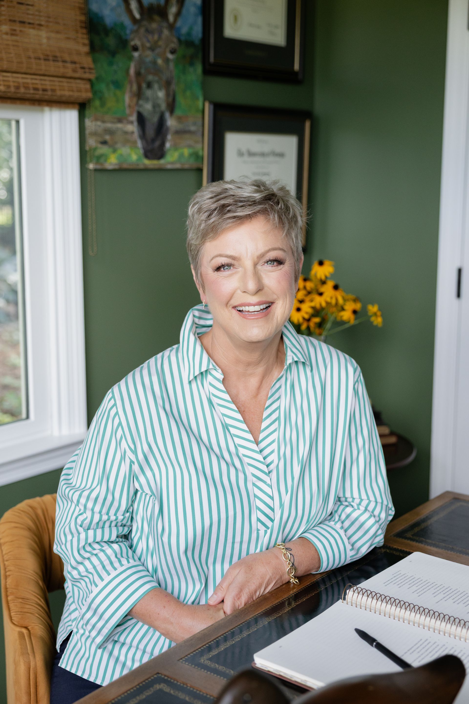 Smiling woman seated at a desk in a green office, wearing a striped shirt with papers and a notebook nearby. Dale Bowers.