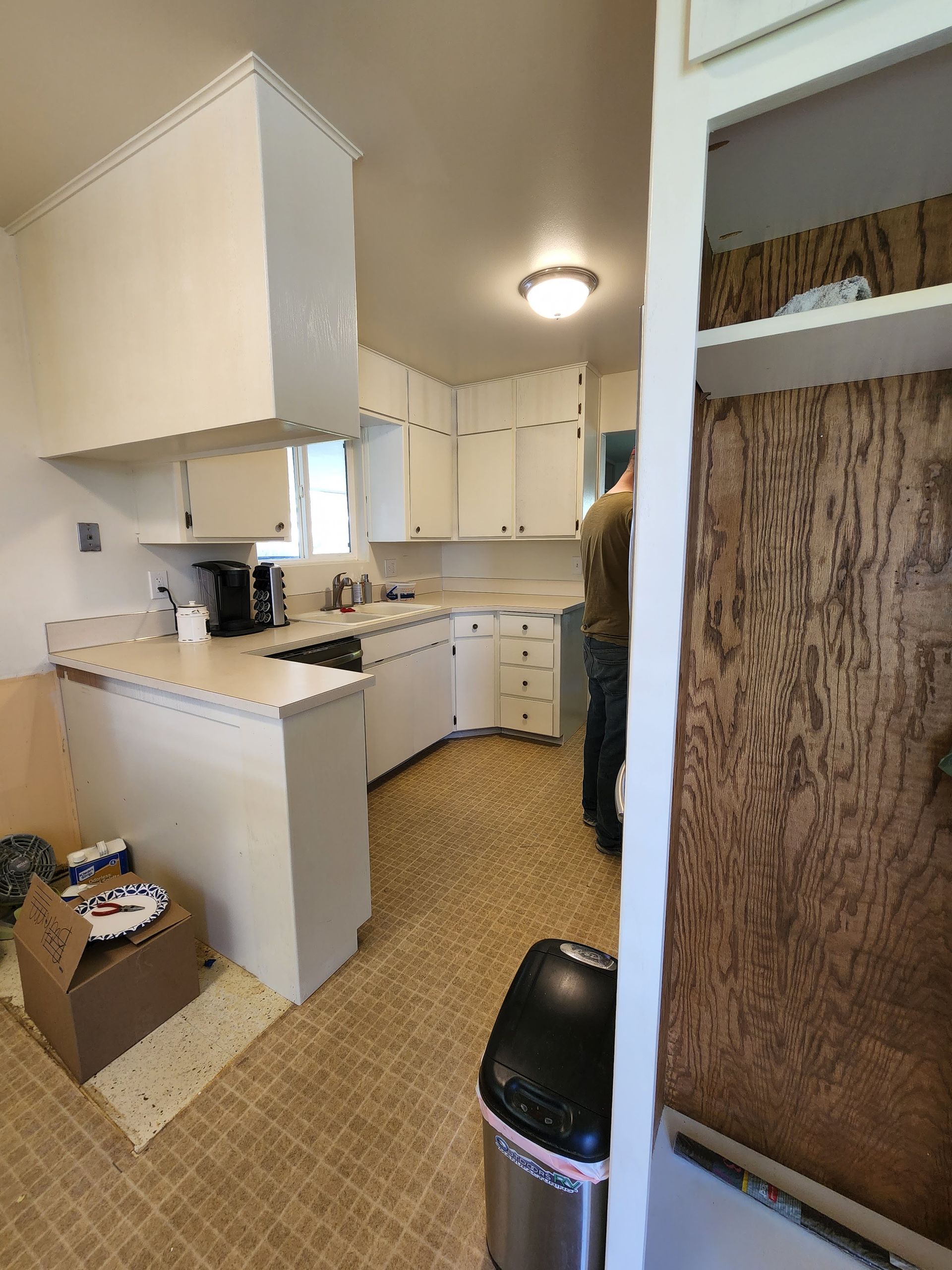 A kitchen with white cabinets, beige counters and floor. A person stands near a pantry.