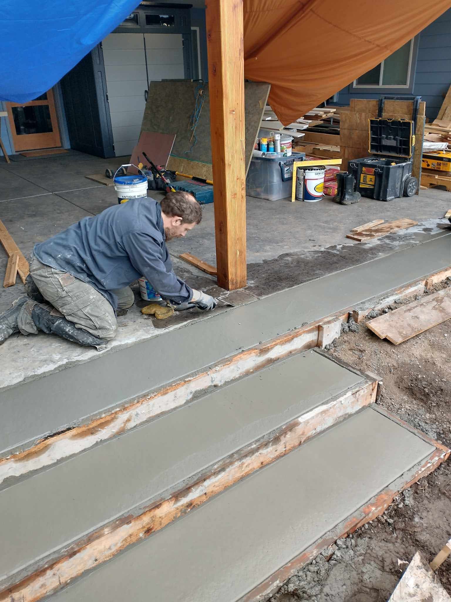 Construction worker smoothing wet concrete steps. Outdoors, under a tarp, near a wooden post.