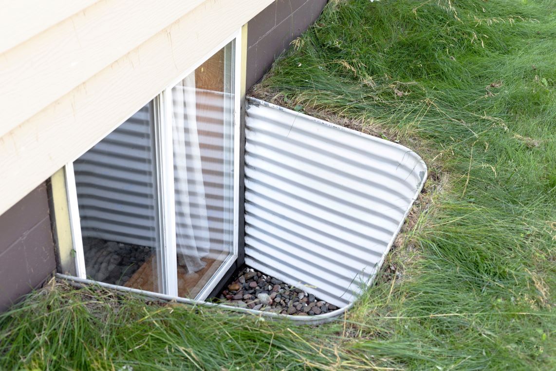 Basement window with corrugated metal well in the side of a house, surrounded by grass.