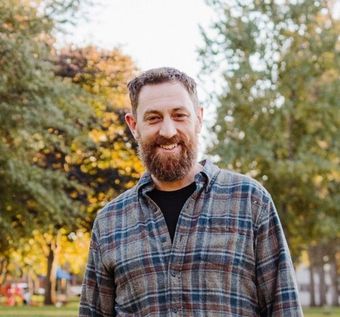 Man with a beard smiling, wearing a plaid shirt, standing outdoors in front of trees with autumn foliage.