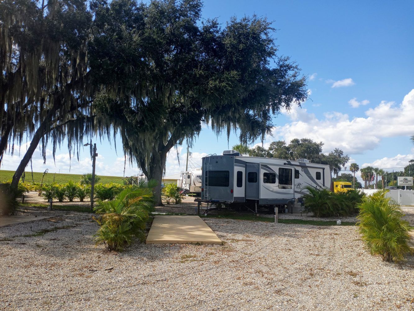 RV parked on gravel in a campground. Green bushes and tree in view, blue sky.