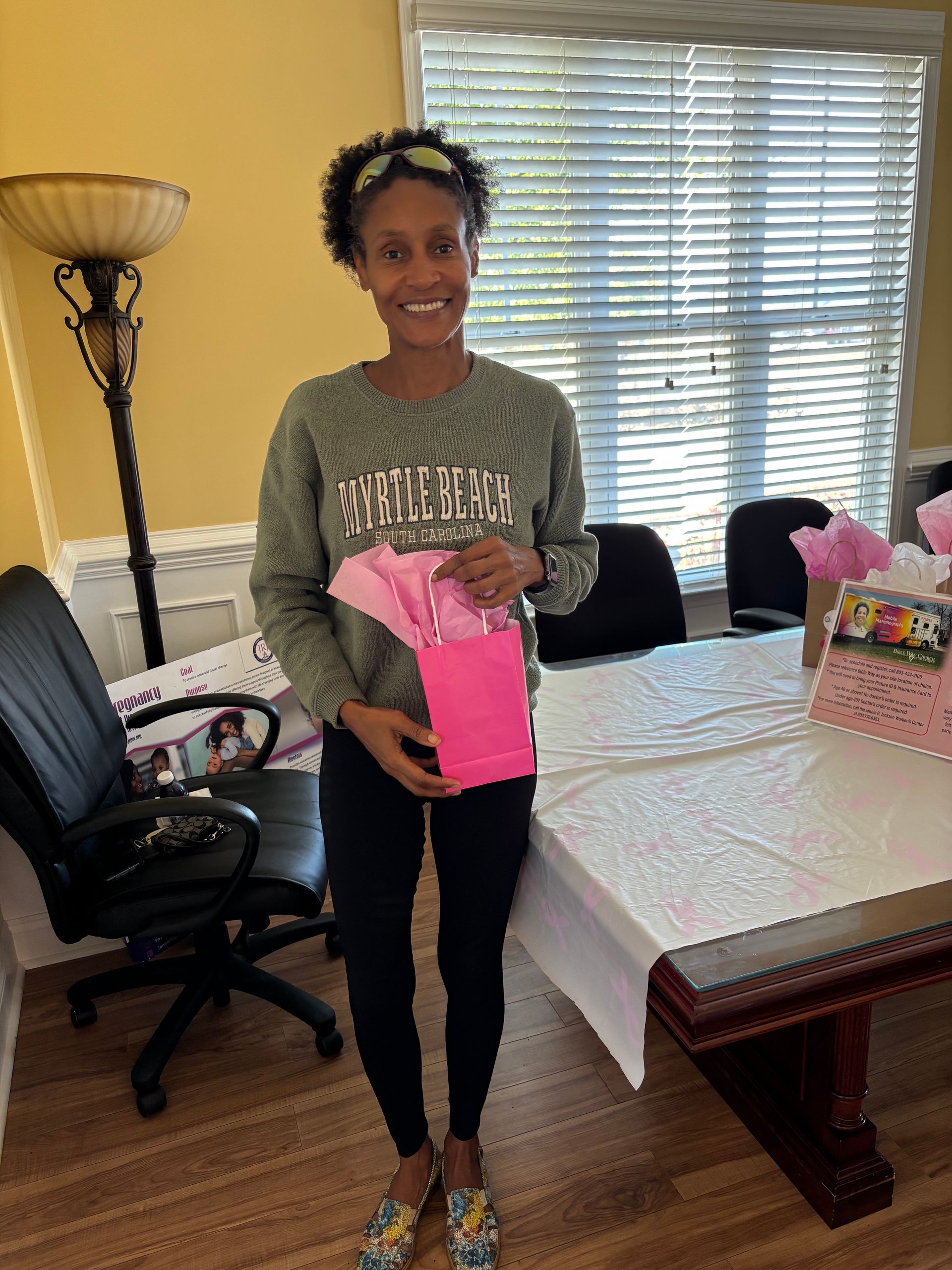 Woman holding a pink gift bag, smiling, standing in front of a table with flowers.