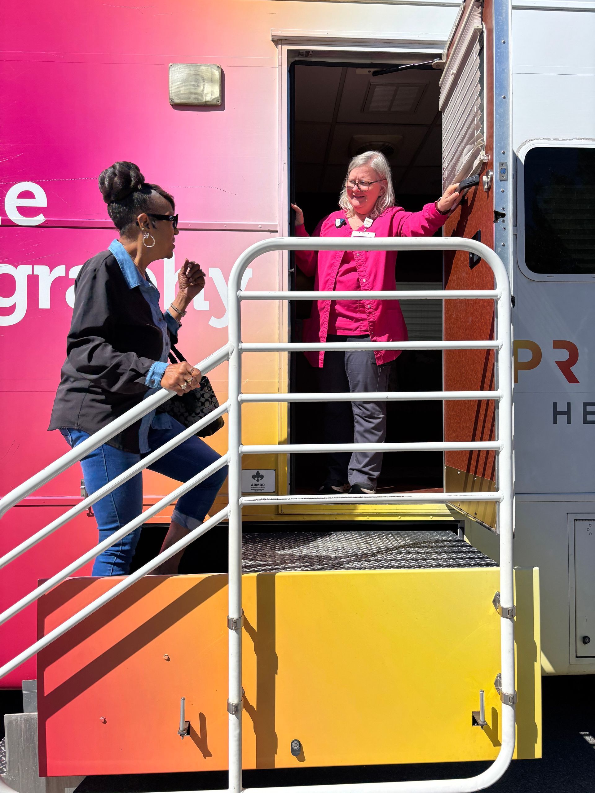 Two people at the entrance of a mobile health clinic, one ascending steps, the other gesturing inside.