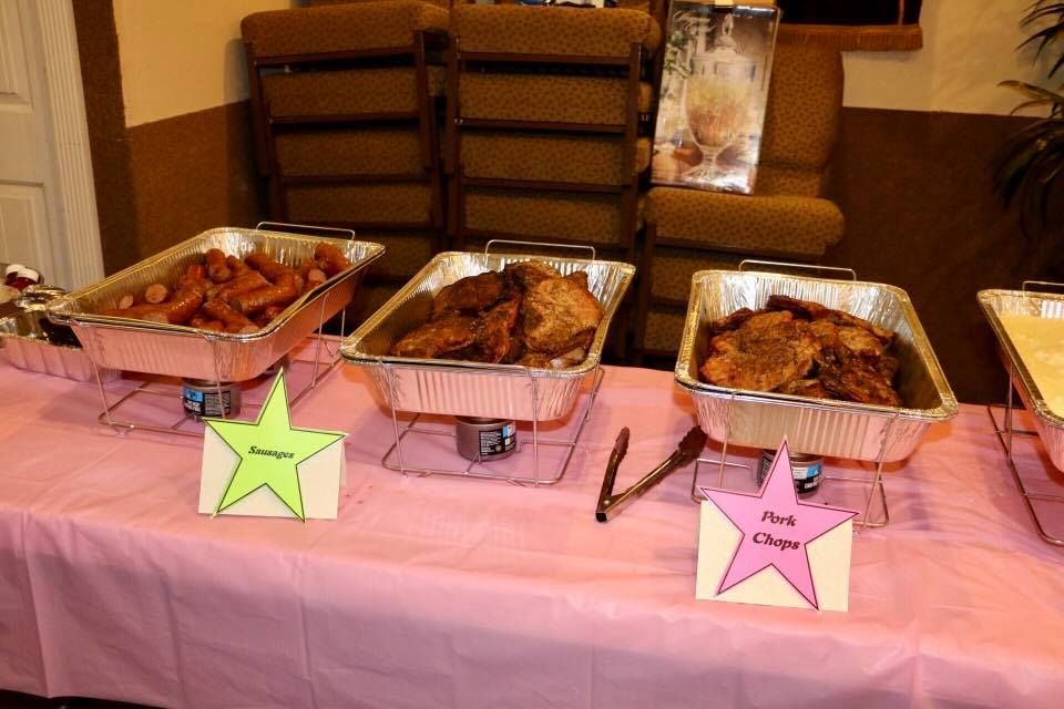 Three trays of roasted food on a pink tablecloth with labeled cards in a banquet setting