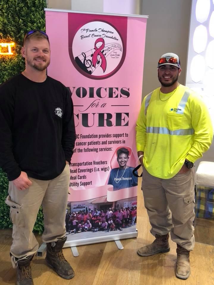 Two men standing beside a pink Voices for the Cure banner indoors