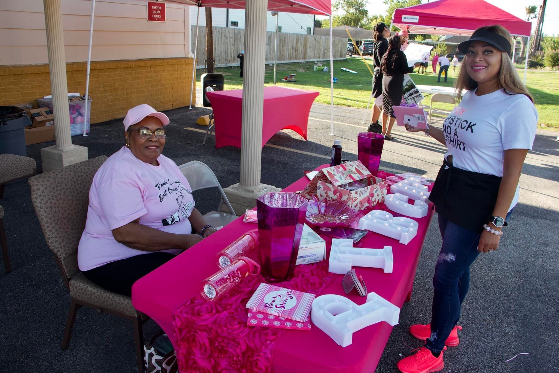Two people at a pink-covered outreach table with brochures and gifts under a tent outdoors