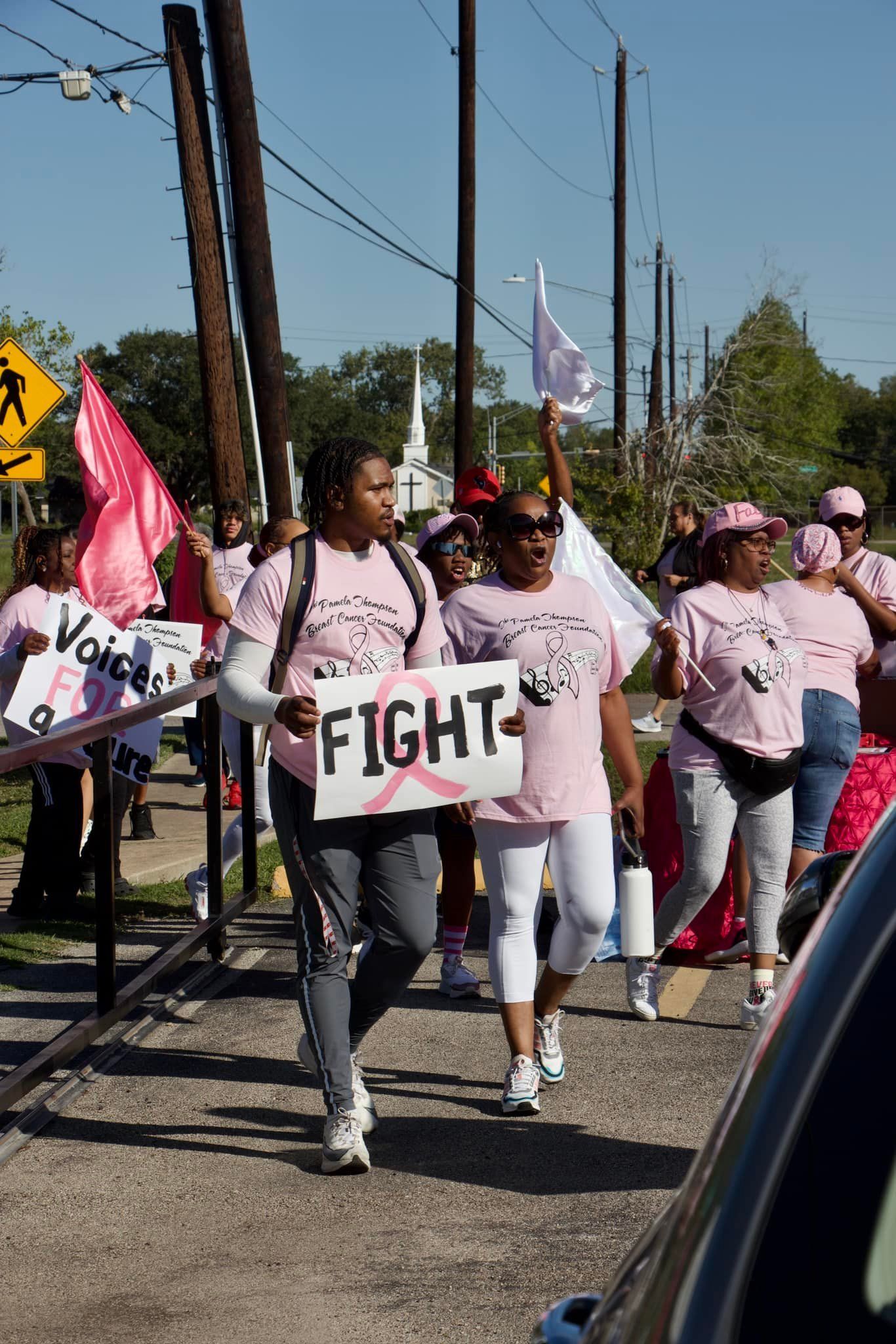 Protesters marching on a street holding signs, including one reading “FIGHT,” and pink flags.