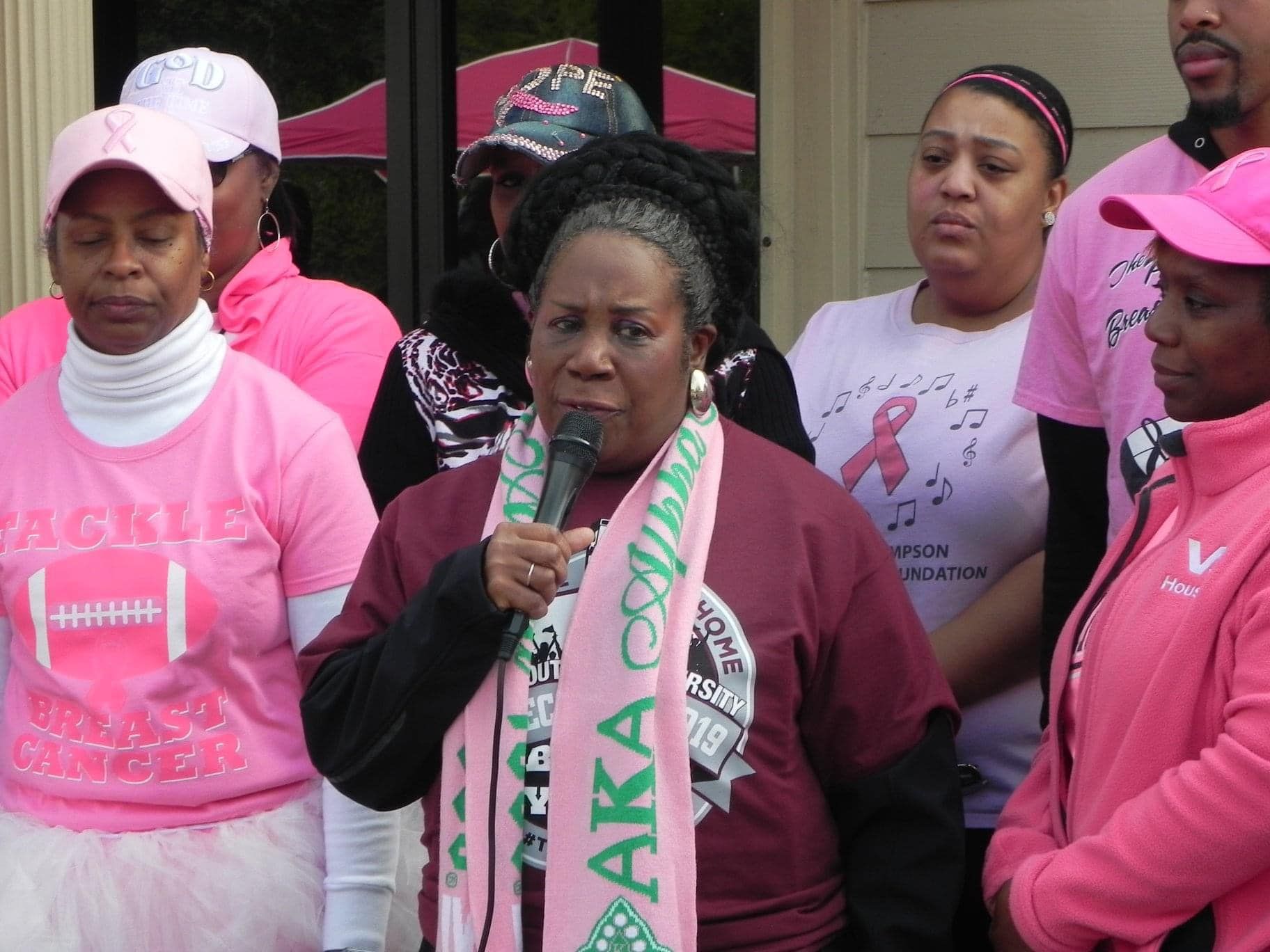 Woman speaking into a microphone at a pink rally, surrounded by supporters in matching shirts and hats