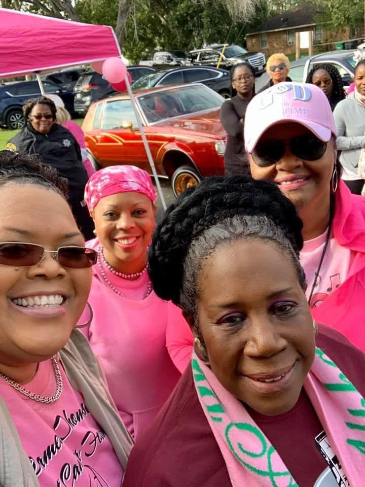Group of smiling people in pink shirts taking a selfie outdoors near parked cars.