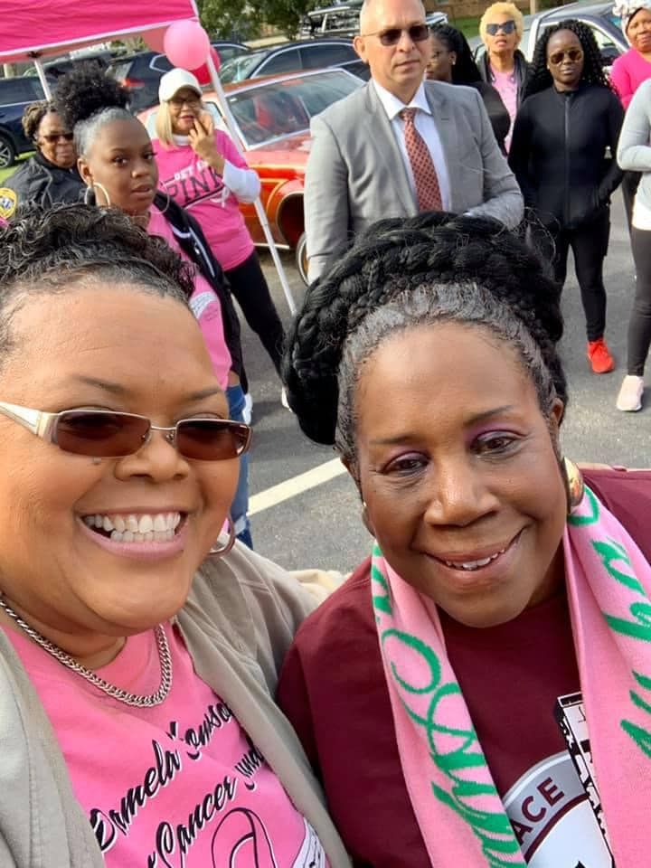 Two smiling women posing outdoors in pink attire at a community event.