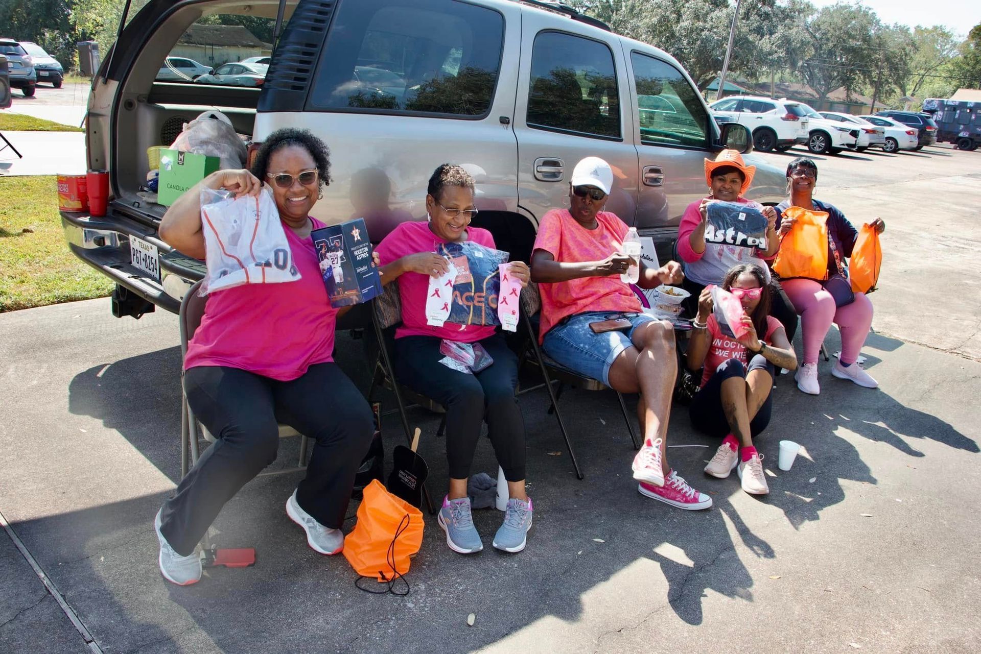 Six people sit on a curb beside an SUV, holding pink items and posing outdoors in a parking lot.