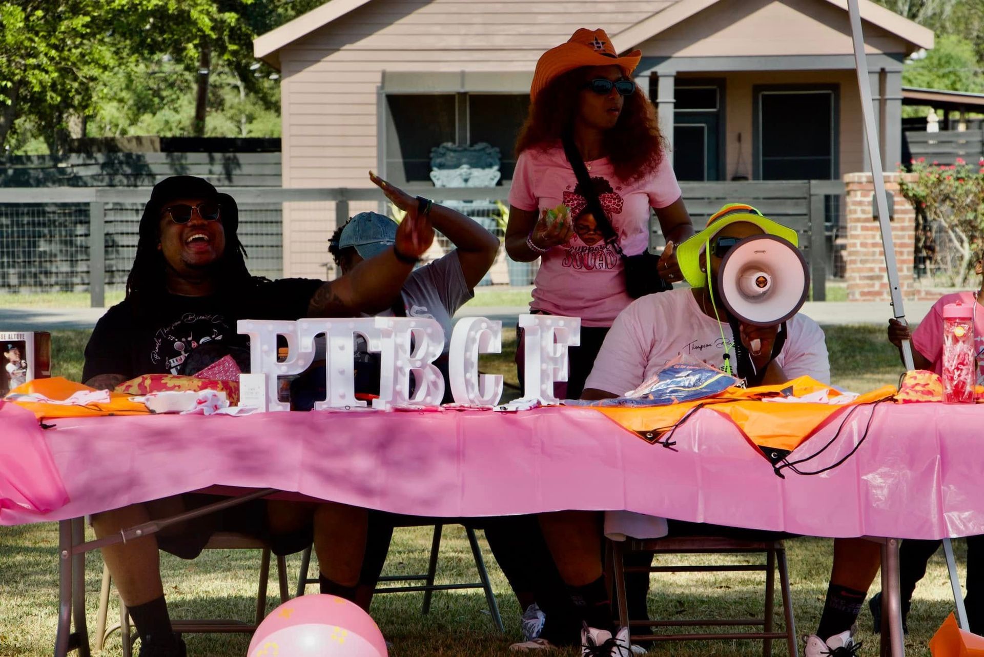 Three people behind a pink table with balloons and large letters, speaking into a megaphone outdoors.