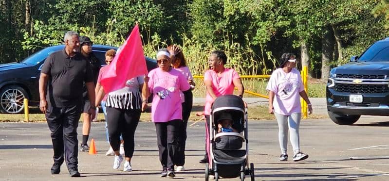 People walking outdoors in a parking lot, one pushing a stroller and another carrying a bright pink flag.