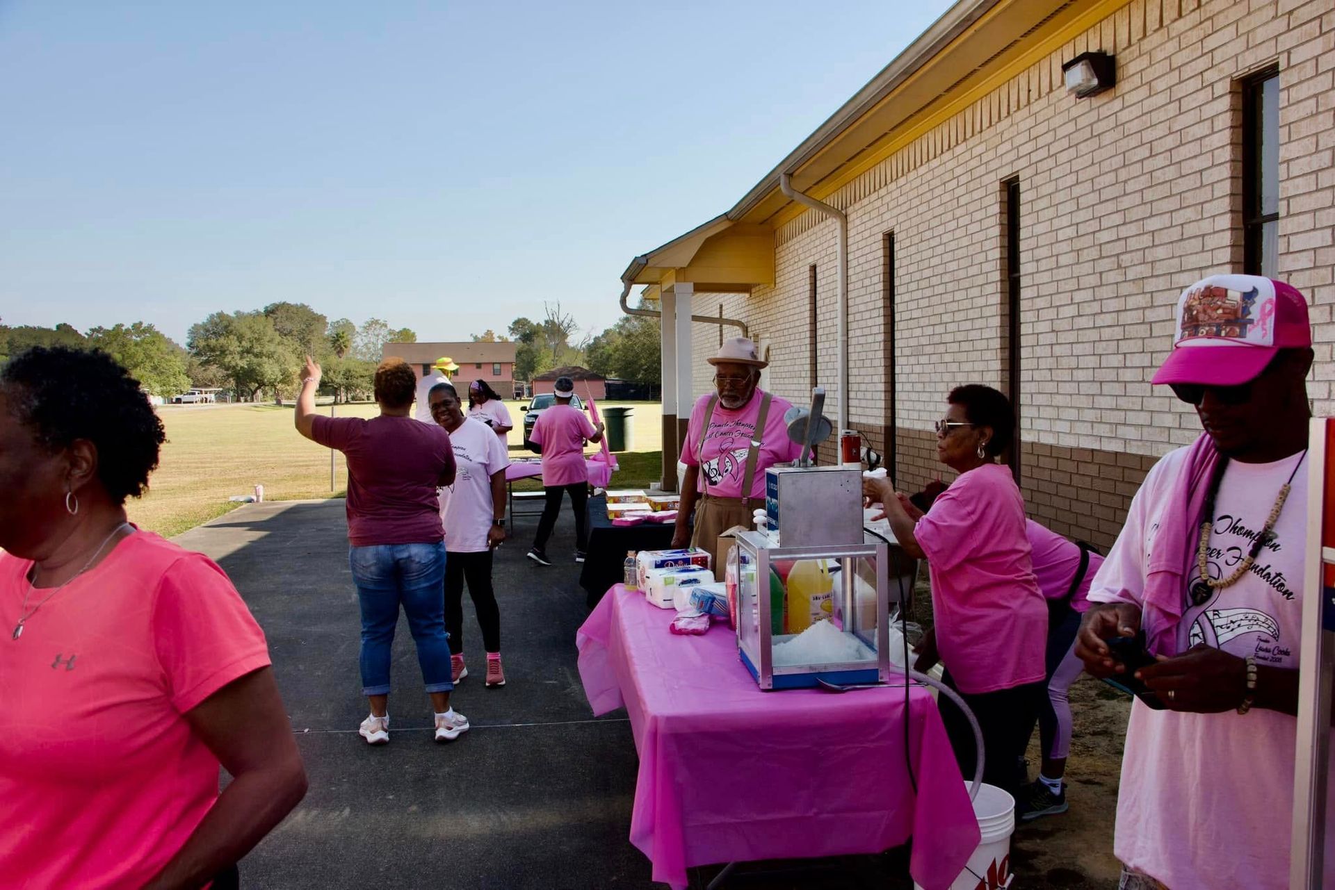 People in pink shirts serve at a pink-draped table outside a brick building.