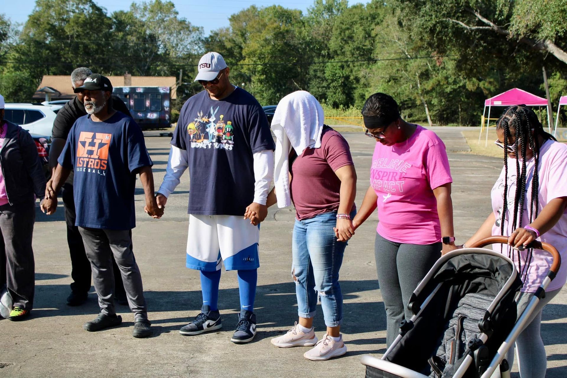 People standing outdoors in a parking lot, some holding hands, with a stroller at right.