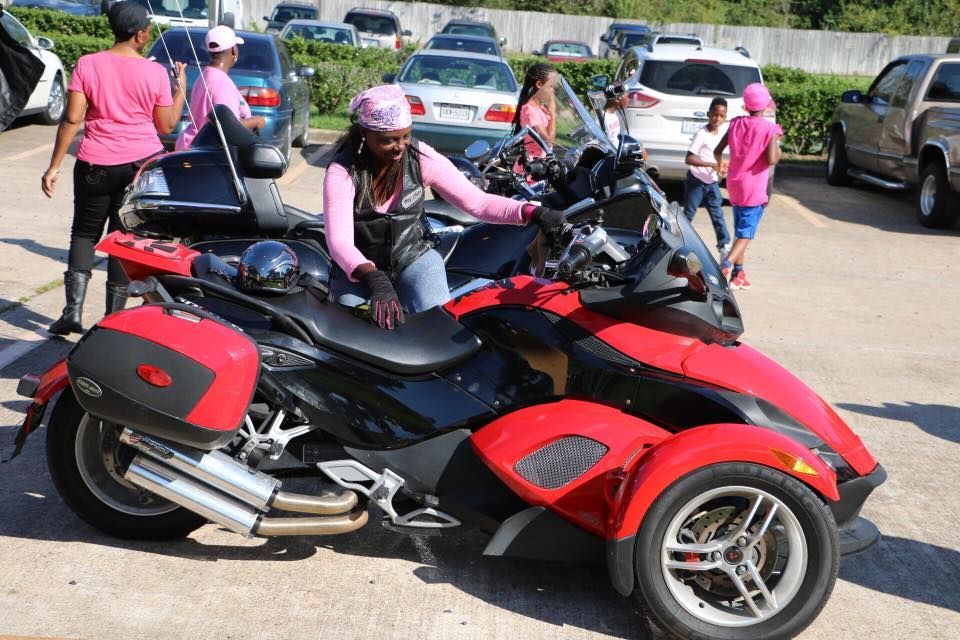 Person riding a red-and-black trike motorcycle in a parking lot, with others and cars nearby