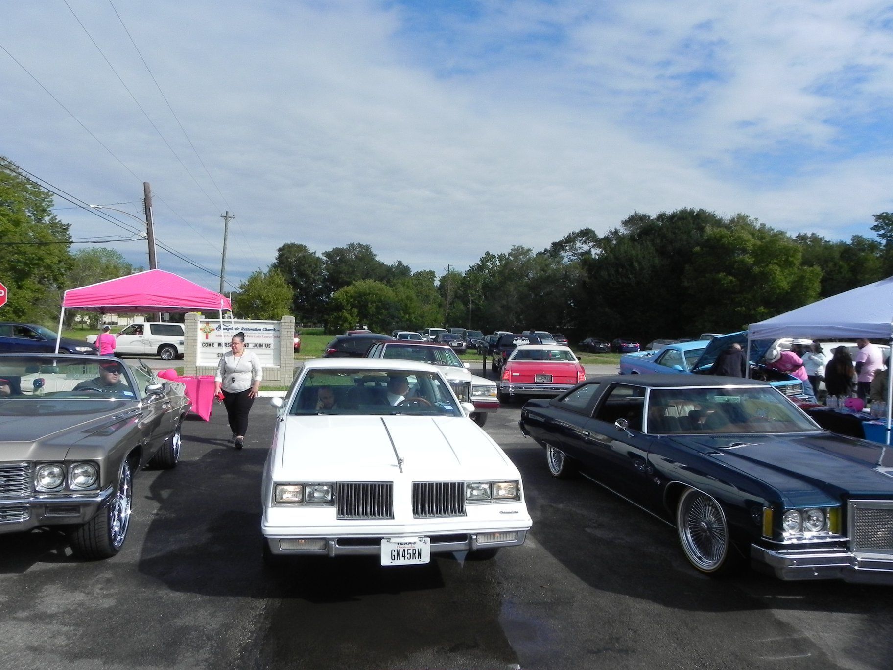 Classic cars parked at an outdoor car show under a blue sky, with tents and spectators nearby.