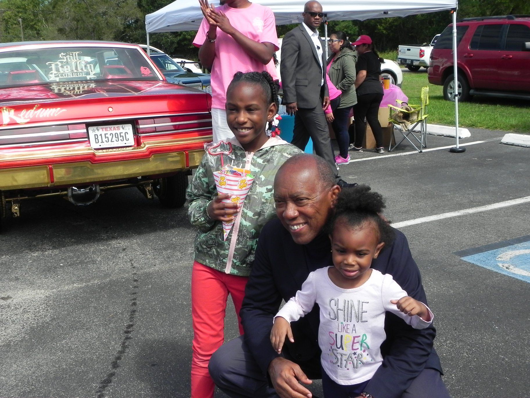 Two children pose with a smiling man in a parking lot beside classic cars and tents.