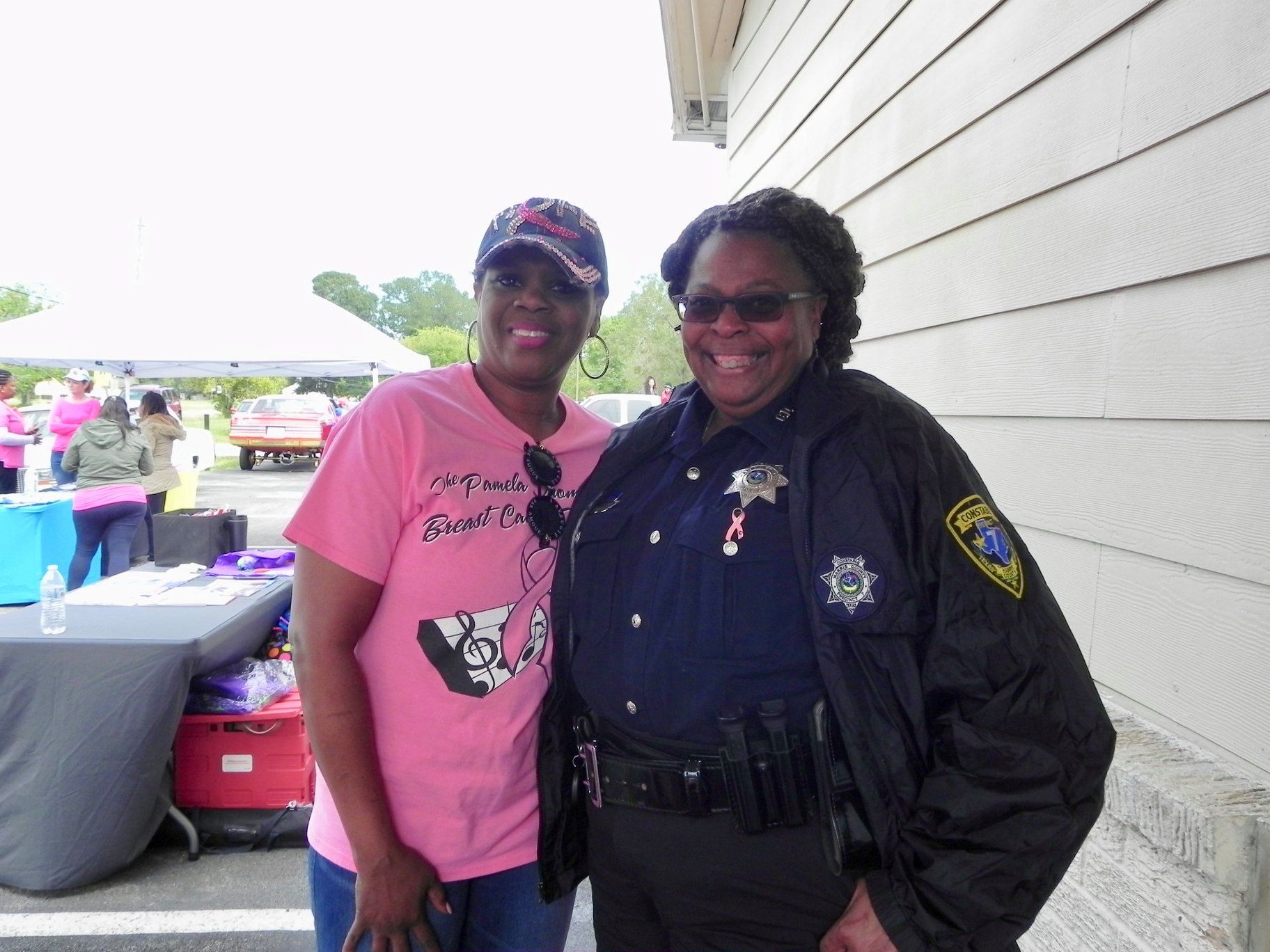 Two smiling people stand beside a white building, one in a pink shirt and one in a dark uniform.