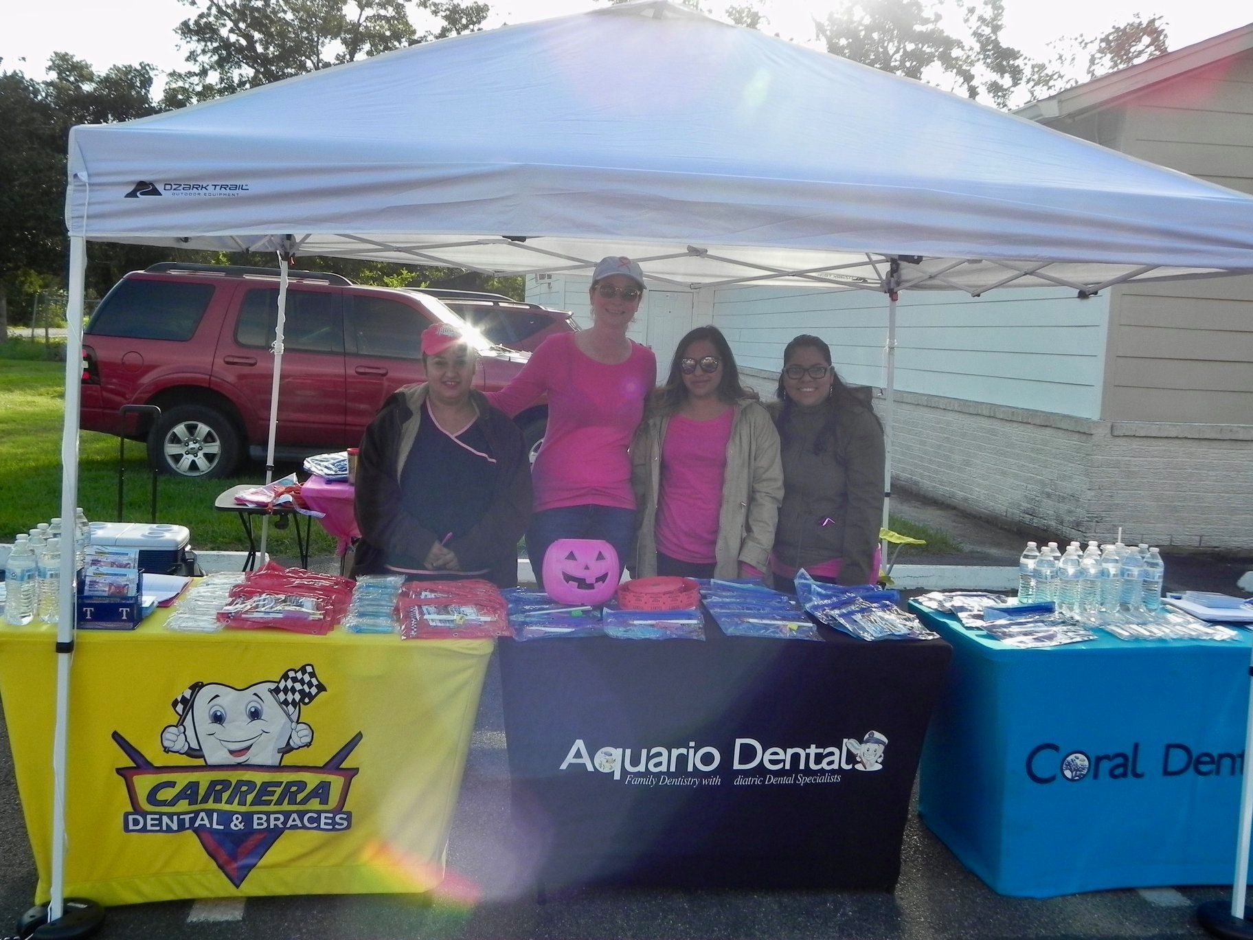 Four people behind a dental outreach table under a white tent, with colorful clinic banners and supplies.