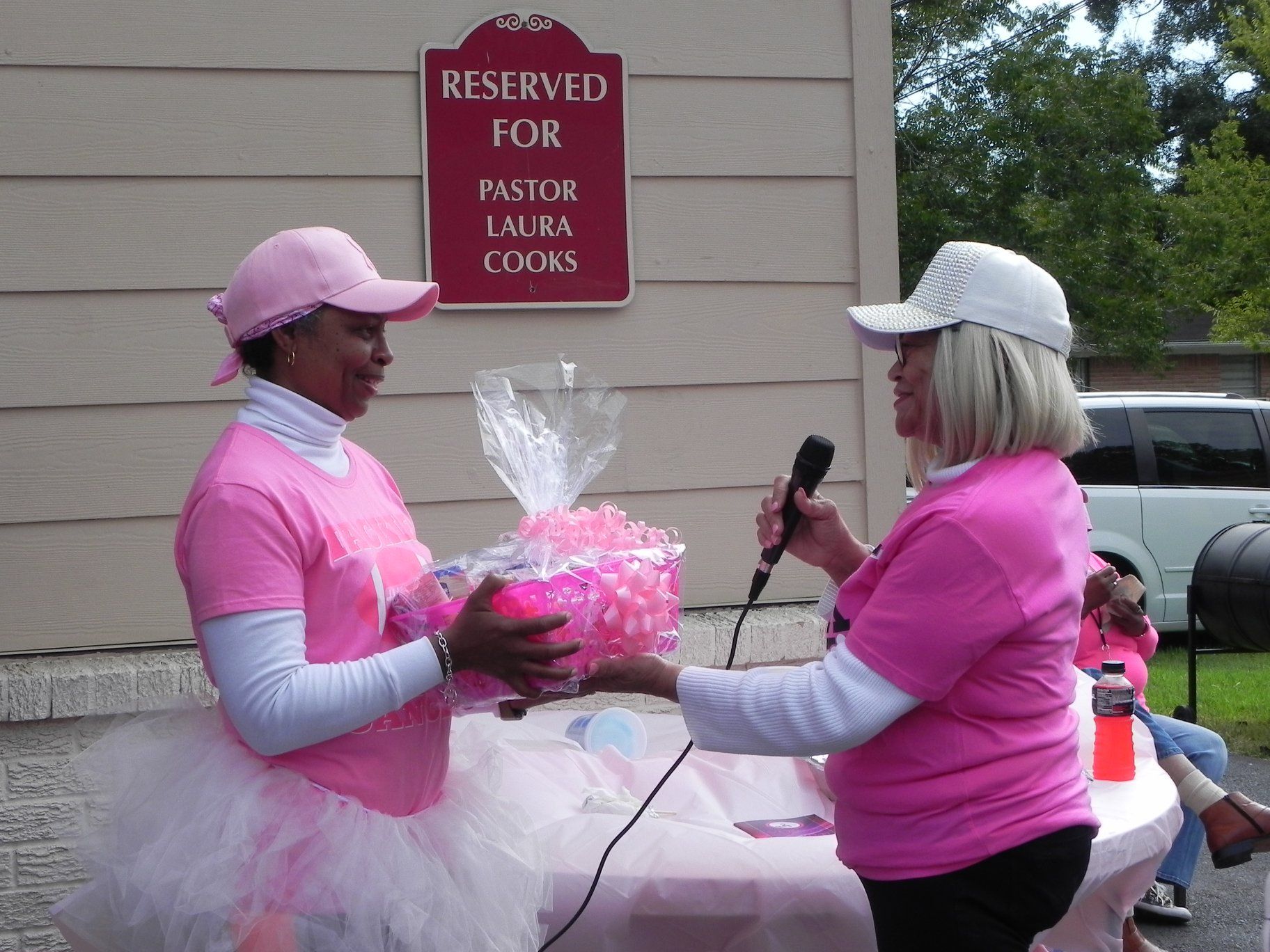 Two women in pink outfits exchange a gift basket beside a reserved sign.