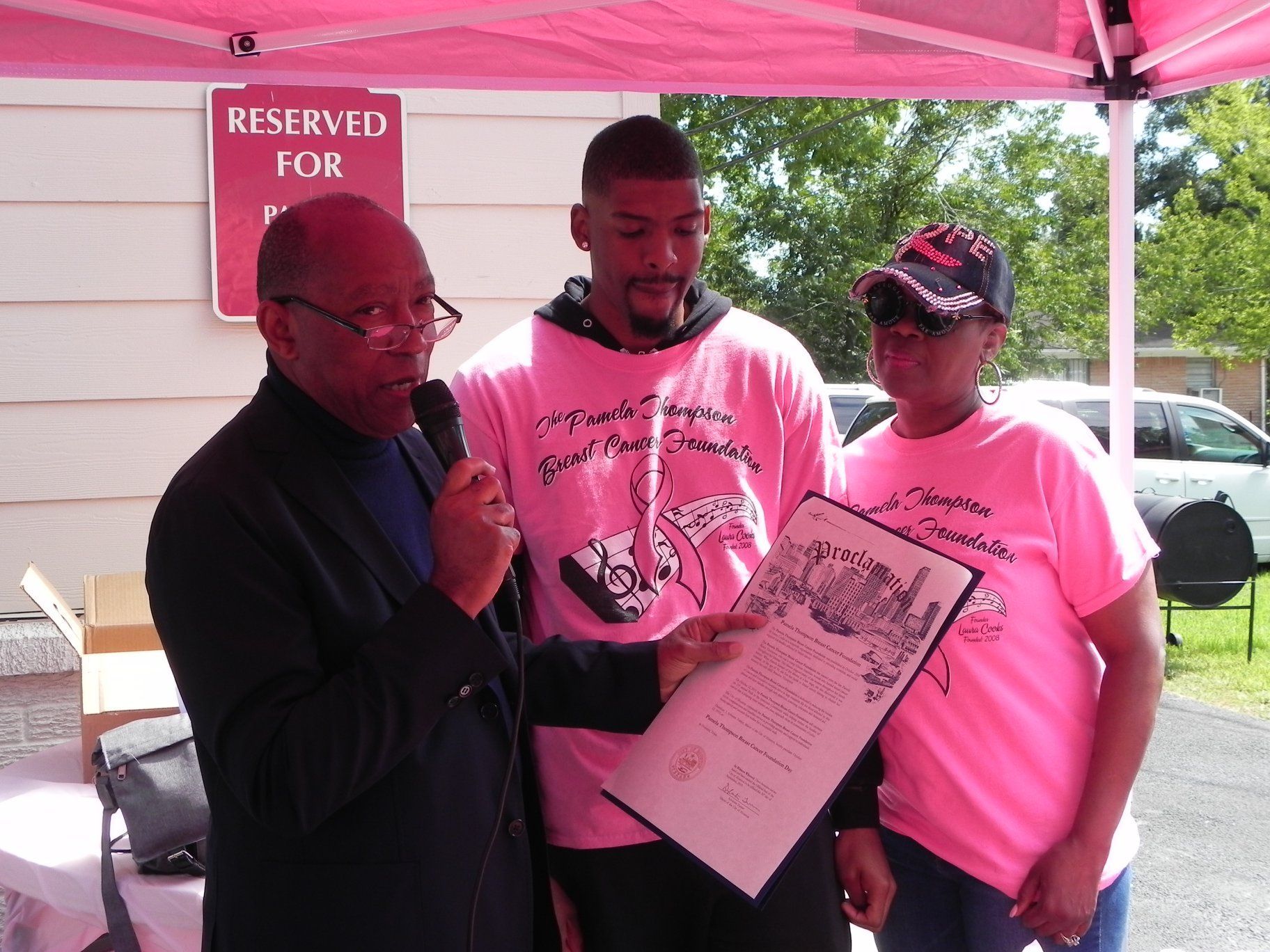 Three people under a pink tent hold a certificate at an outdoor event.