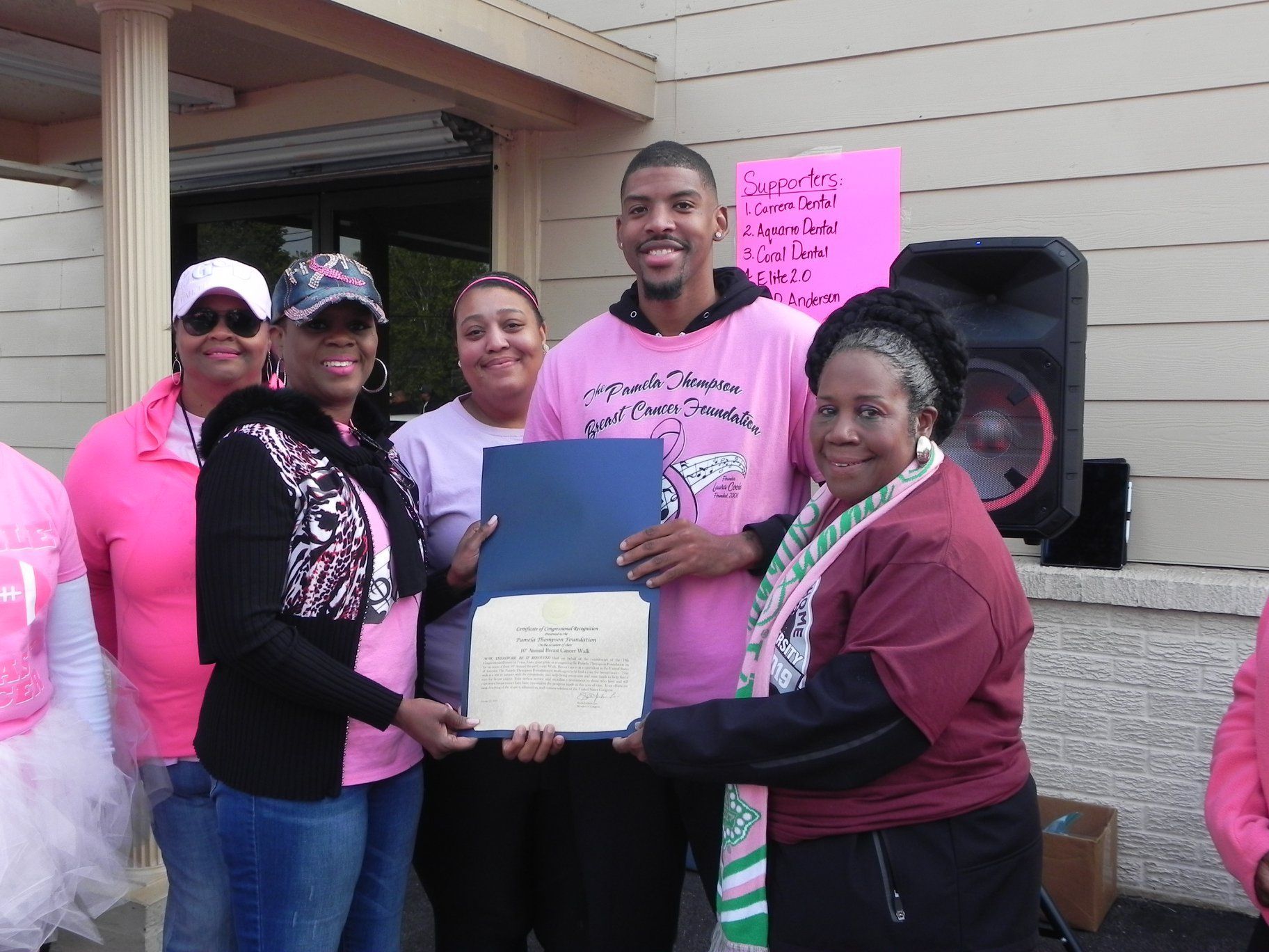 Group celebrating outside a building, holding a blue folder and certificate beside a pink sign and speaker