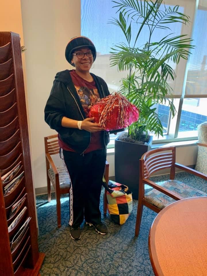 Person holding a red gift basket in a bright indoor room with potted plants and chairs.