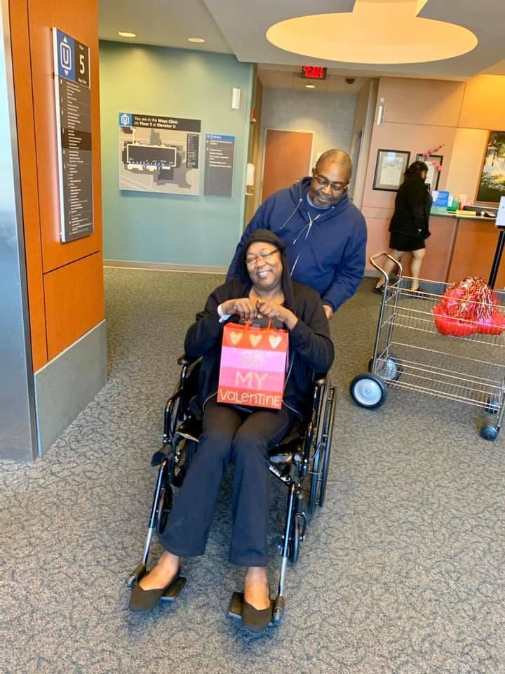 Two people in a wheelchair at a hospital entrance, one holding a pink sign and one pushing from behind.