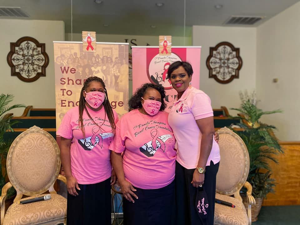 Three people in pink shirts and masks, standing in a church, in front of pink banners.