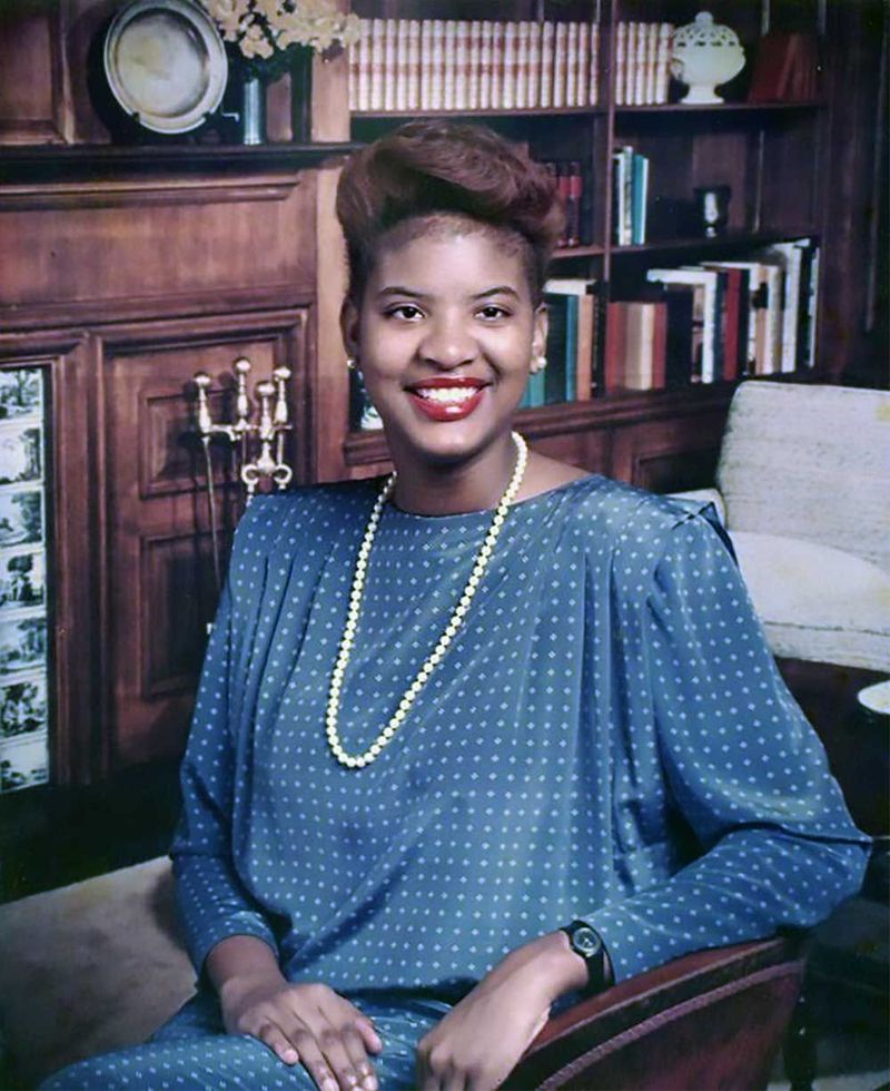 Person seated in a chair, smiling in a wood-paneled room with bookshelves and a fireplace.