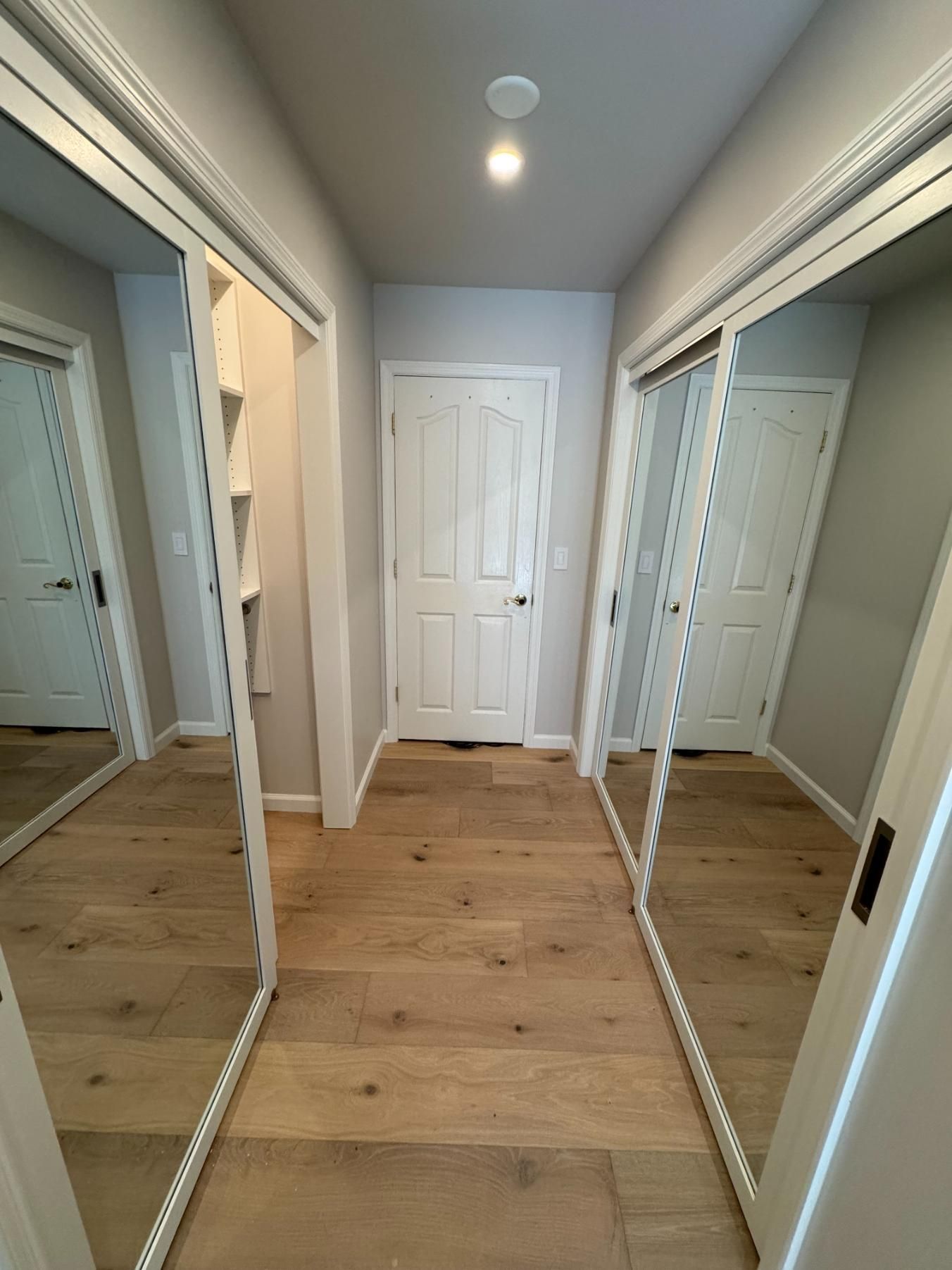 Narrow hallway with mirrored closet doors, light wood floor, and a white door at the end.