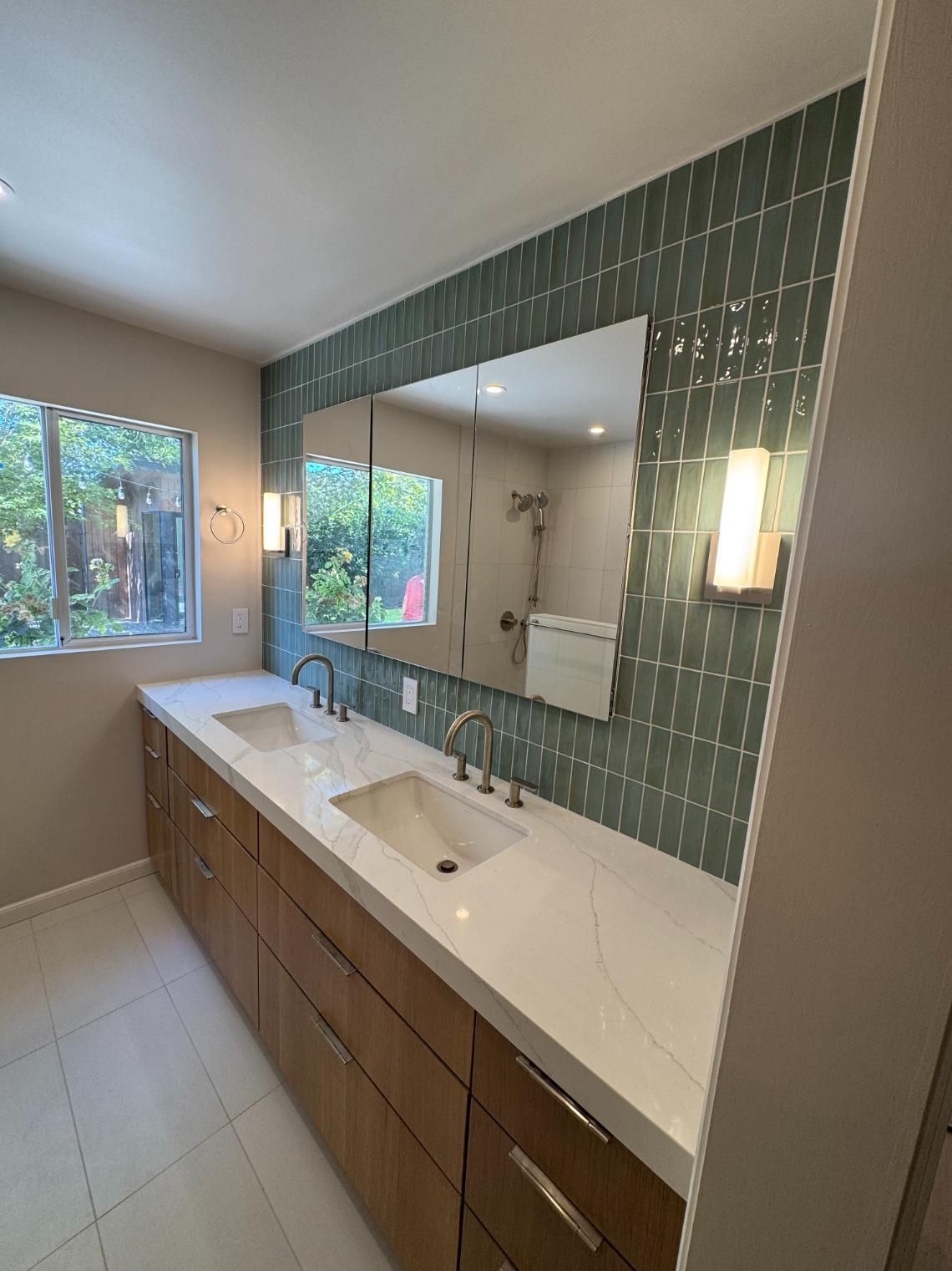 Modern bathroom vanity with double sinks, large mirror, green tile backsplash, and window view of trees