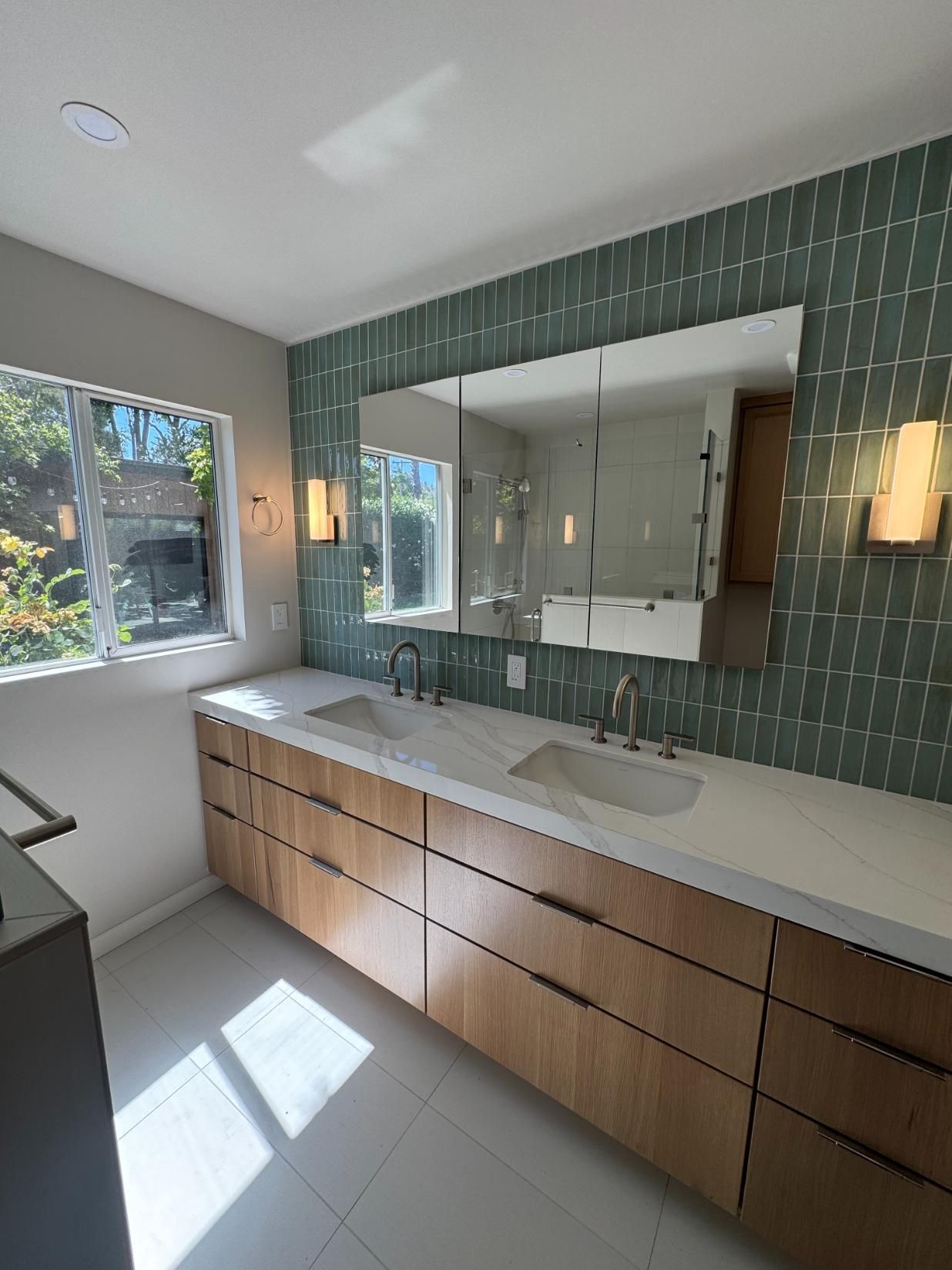 Modern bathroom vanity with green tile wall, double sinks, large mirror, and natural light from windows