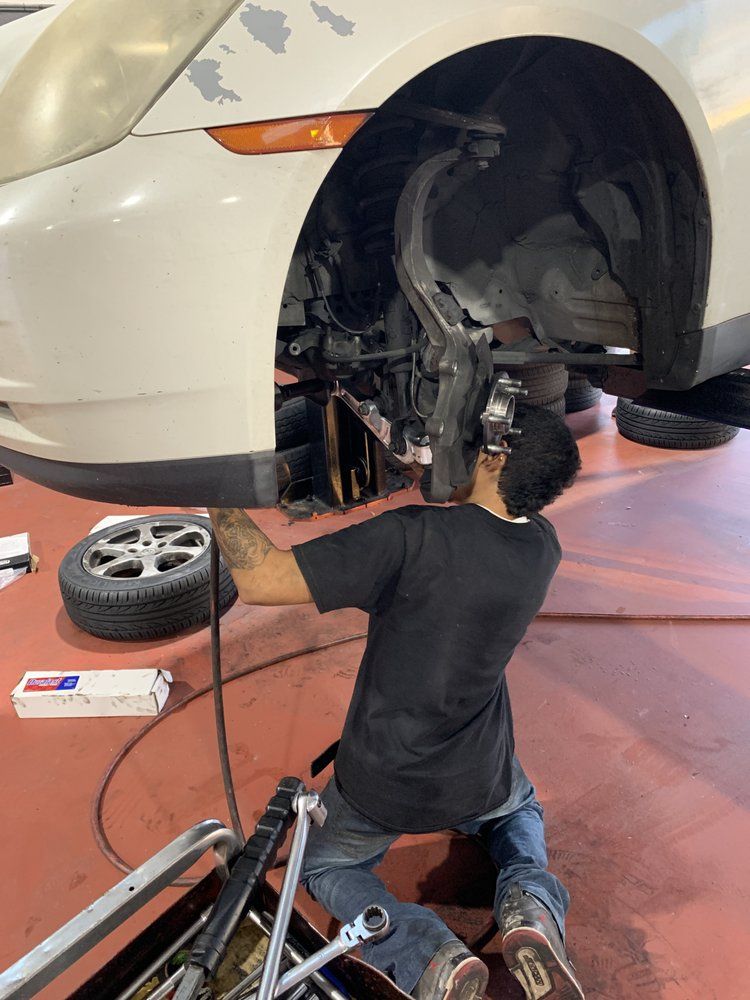 Mechanic working under a raised white car beside a removed wheel in a garage | Elite Auto Repair & Service Inc