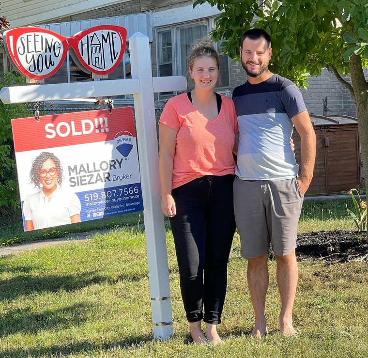 Smiling couple standing beside a sold real estate sign in front of a house
