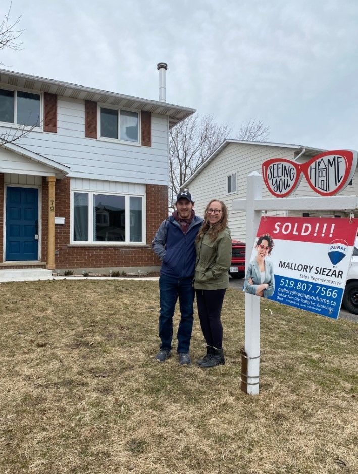 Two people posing beside a sold real estate sign in front of a house.