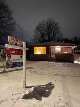 Snowy nighttime real estate sign with a sold notice in front of a lit house
