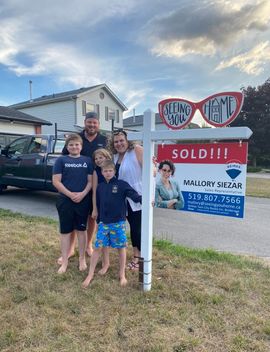 Family posing beside a sold real estate sign in front of a house at dusk