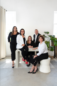 Five people posing in a bright studio, seated and standing around a round table with plants nearby