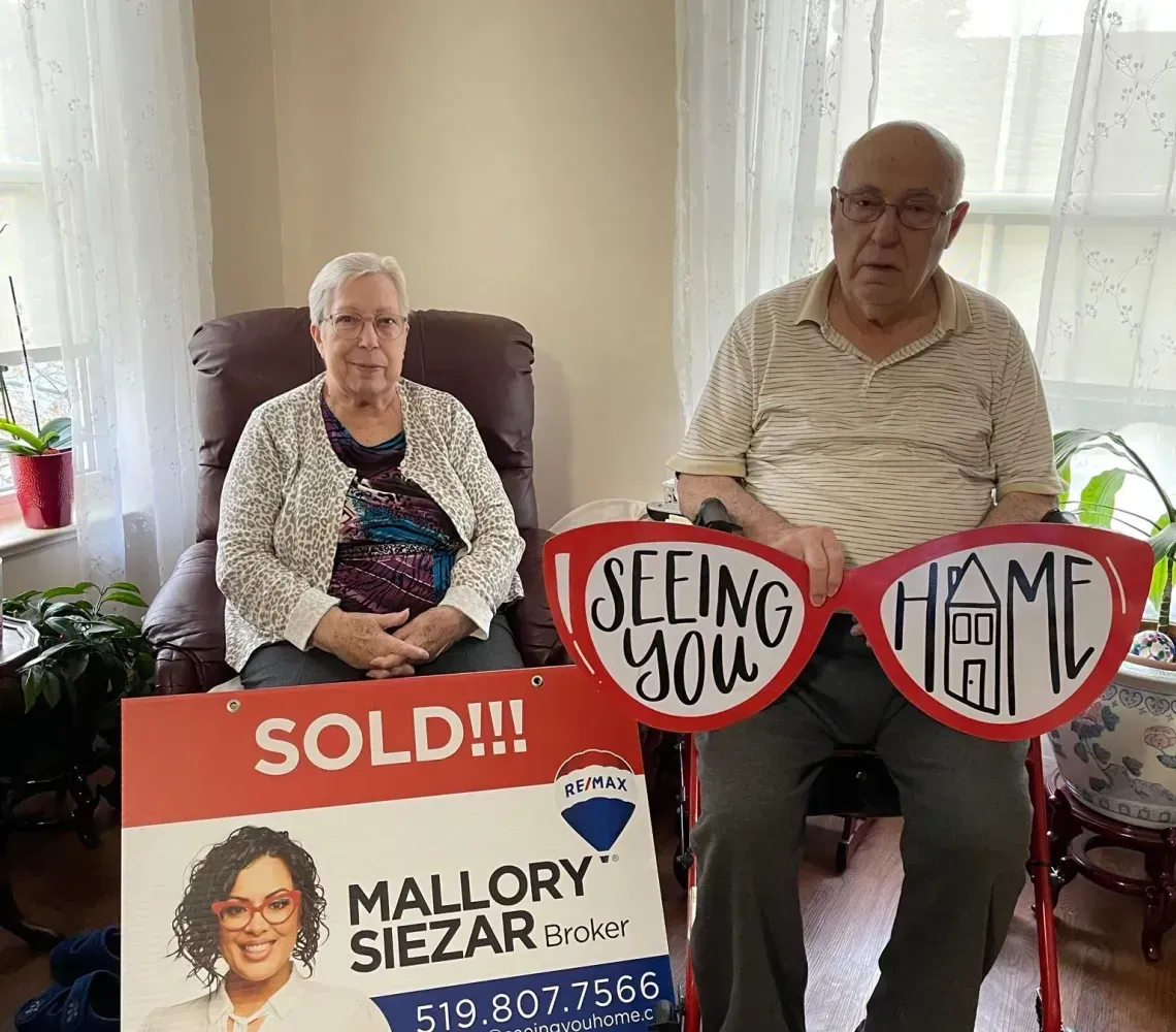 Two seated people hold campaign signs reading “Sold!” and “Keep Them Home” in a bright room