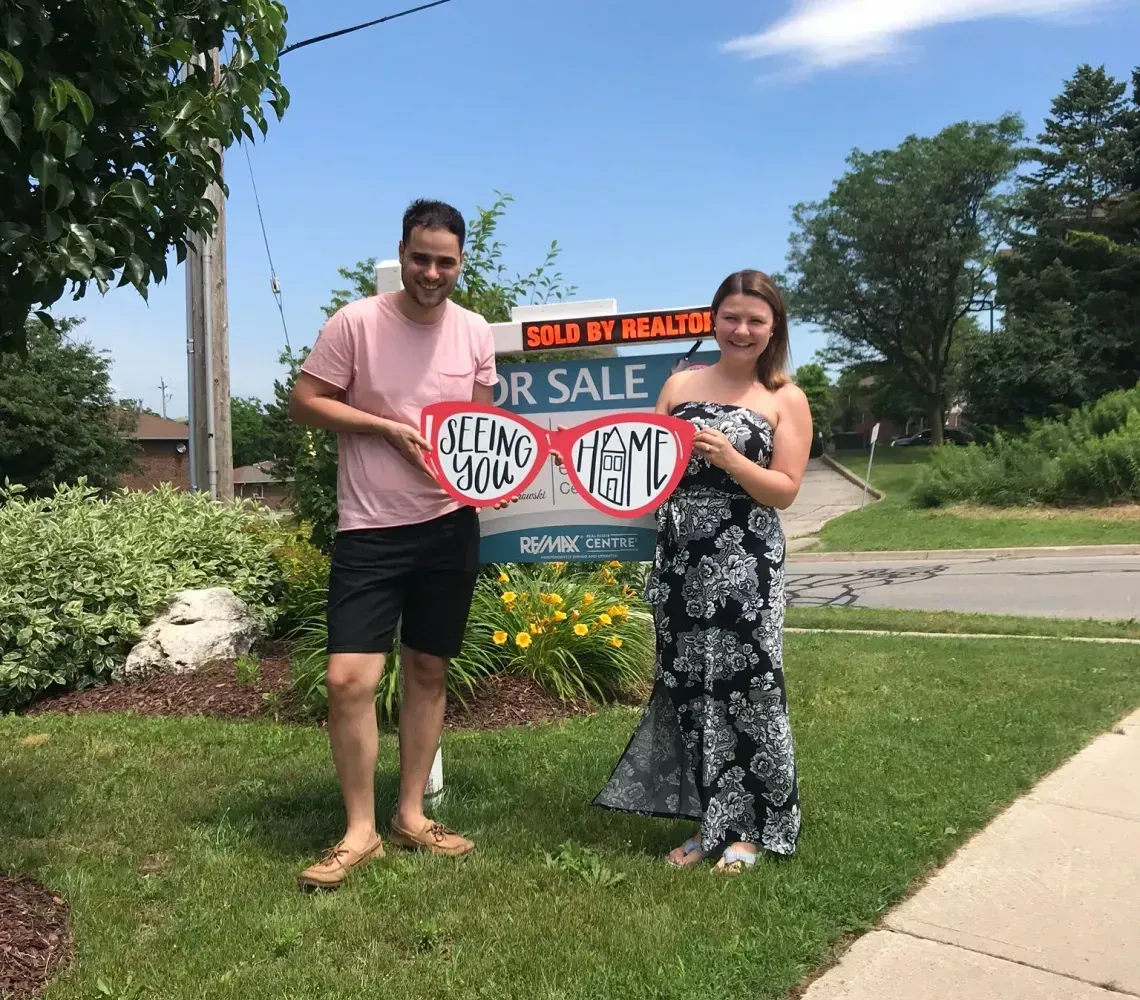 Two people hold a “For Sale” sign on a grassy roadside under a bright blue sky.