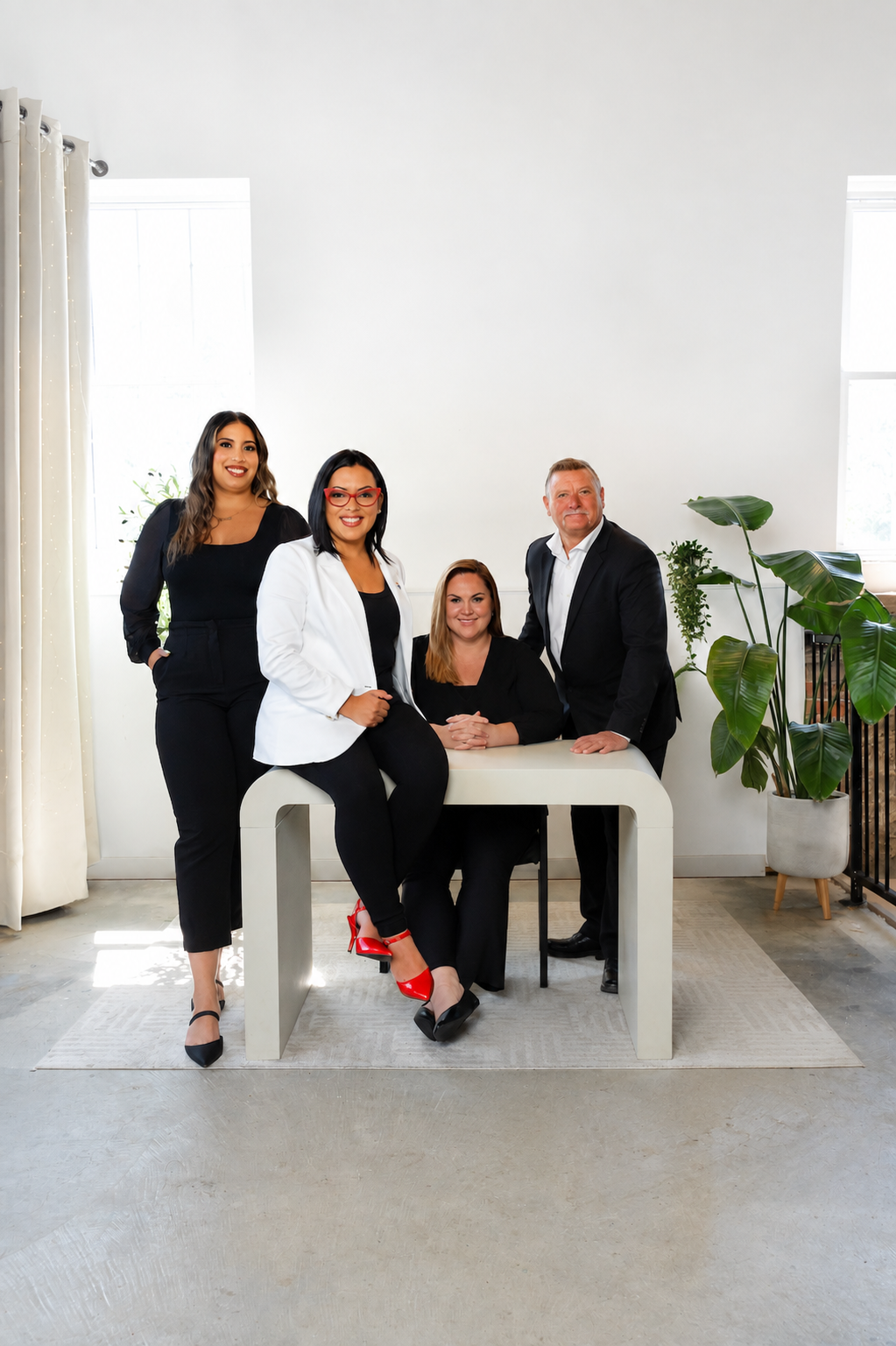 Five people posing in a bright studio, seated and standing around a round table with plants nearby