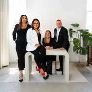 Five people posing in a bright studio, seated and standing around a round table with plants nearby