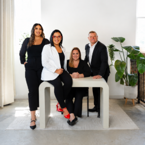 Five people posing in a bright studio, seated and standing around a round table with plants nearby