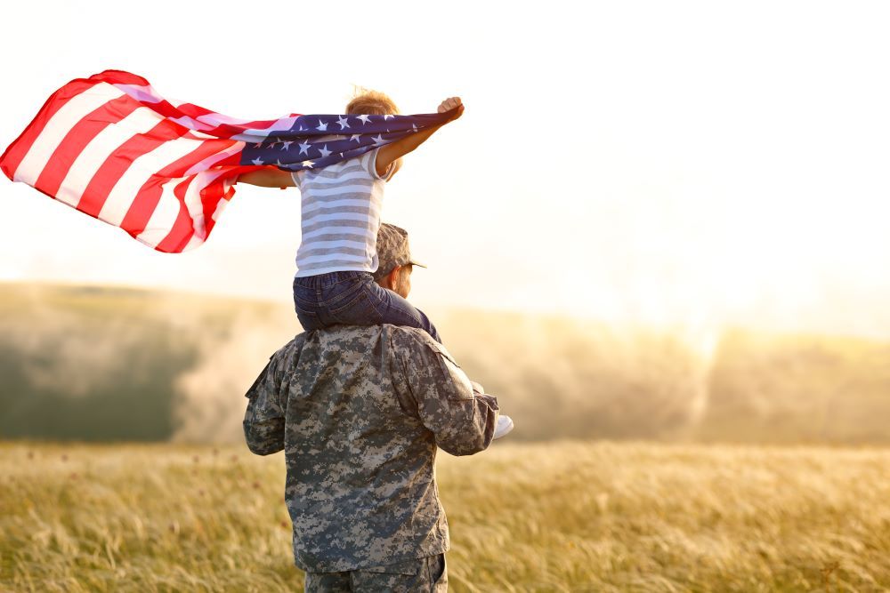 A man in a military uniform is carrying a child on his shoulders with an american flag.